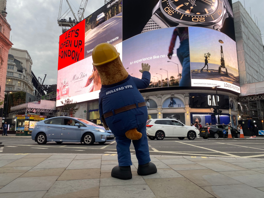Mullvad VPN mole mascot waving at a Piccadilly Circus billboard reading LET'S OPEN UP LONDON. Mascot's blue suit reads MULLVAD VPN.