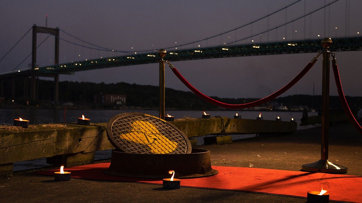 Manhole cover with the Mullvad VPN mole logo and text on a red carpet at night, surrounded by candles with a lit bridge in the background.